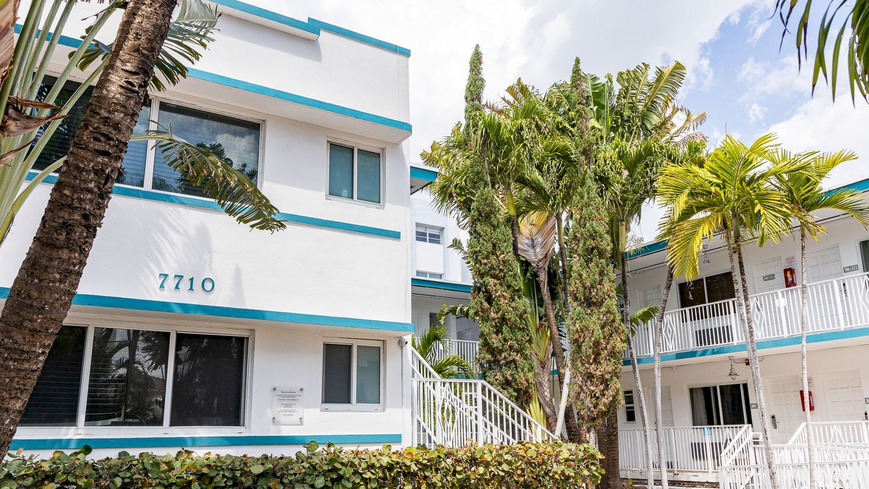 A white and blue apartment building with lush greenery, palm trees, and outdoor seating under a clear sky.