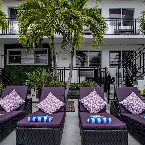 A resort pool area with six black wicker loungers, pink cushions, striped towels, palms, and a two-story white building in the background.