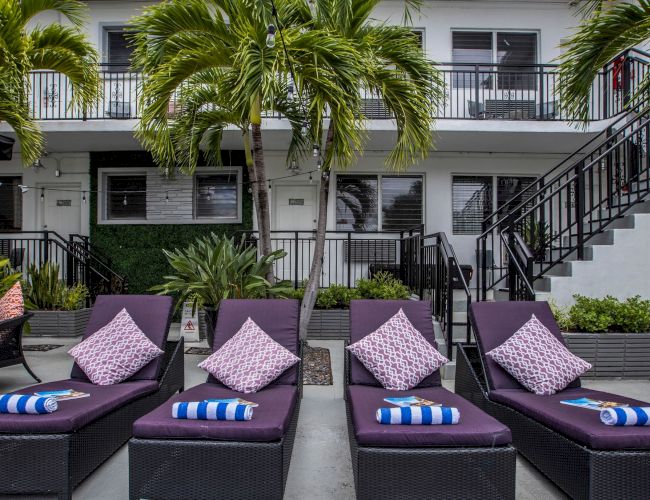 A resort pool area with six black wicker loungers, pink cushions, striped towels, palms, and a two-story white building in the background.