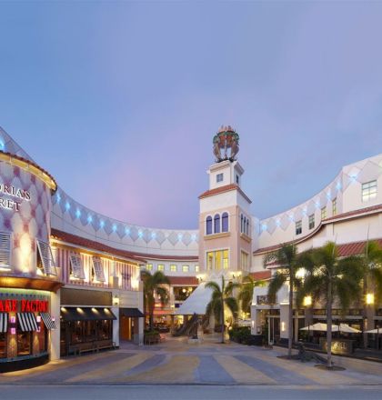 A bright shopping center with palm trees, modern storefronts, and a curved, arcade-style facade; features include a clock tower and Zara store, evening lighting.