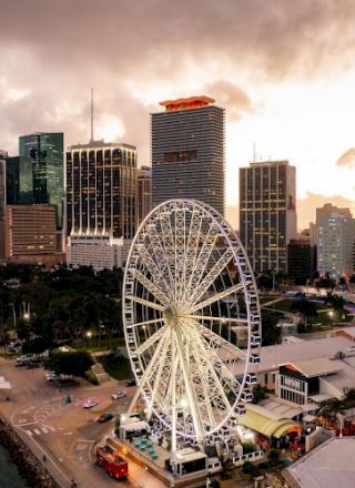 Aerial view of a city skyline with a large ferris wheel by the waterfront, boats docked, and sunset clouds overhead.