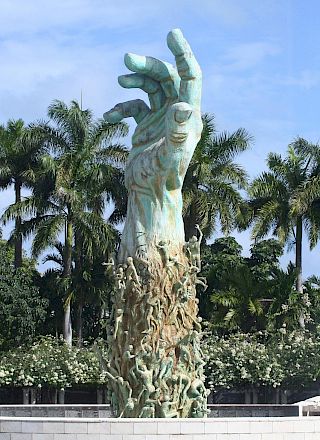 A statue of a robed figure with outstretched arms stands among palm trees under a bright blue sky.