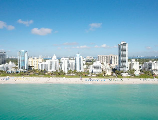 A sunny coastal city skyline with modern high-rises along a turquoise bay and a sandy beach in the foreground.
