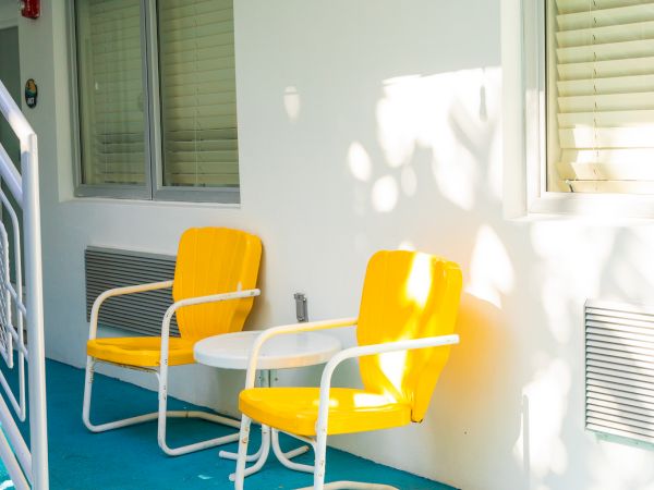 Two yellow chairs and a white table are on a turquoise-painted patio, with sunlight casting shadows on the white wall behind them.