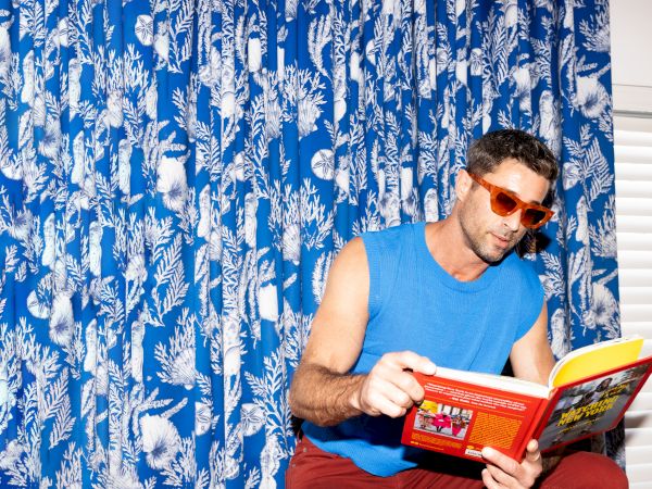 A person wearing sunglasses and a blue sleeveless shirt sits reading a colorful book in front of a blue patterned curtain.