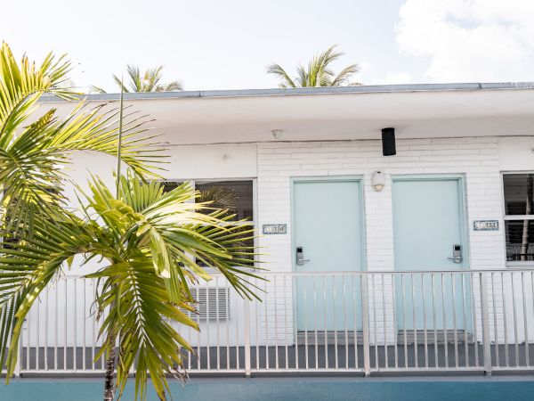 A white building with two light blue doors, a railing, and palm trees in front; partly cloudy sky above.