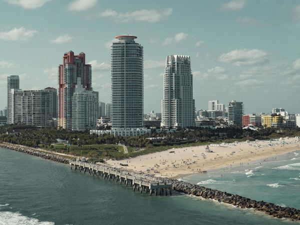 A beach scene with high-rise buildings in the background, a pier stretching into the ocean, and people on the sandy shore under a clear sky.