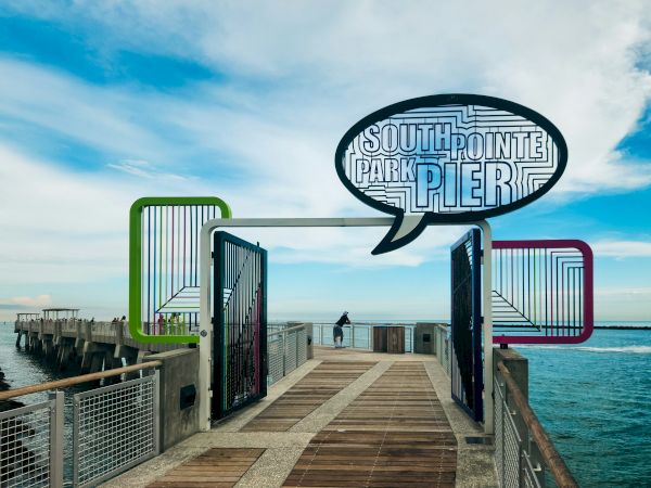 A wooden walkway leads to South Pointe Park Pier, with colorful frames and a speech bubble sign, set against the ocean and blue sky.