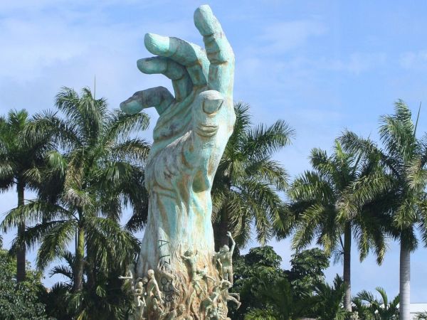 A large hand sculpture with figures climbing its surface, surrounded by palm trees under a blue sky.