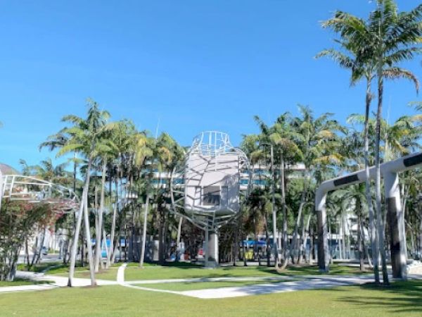The image shows a park with palm trees, modern sculptures, and a clear blue sky. The scene is tranquil and landscaped.