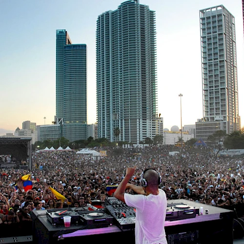 A DJ performs on stage in front of a massive crowd, with tall city buildings in the background at sunset during a lively outdoor concert.