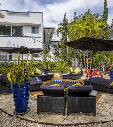A seating area with black wicker furniture, blue and yellow pillows, and large potted plants, in front of a white building.