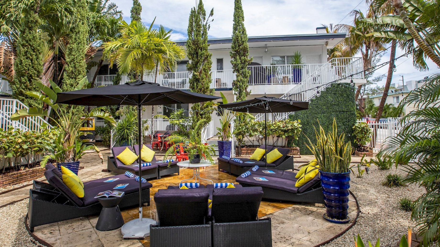 A stylish outdoor lounge area with black wicker seating, yellow cushions, shaded by umbrellas, surrounded by tropical plants and a modern white building.