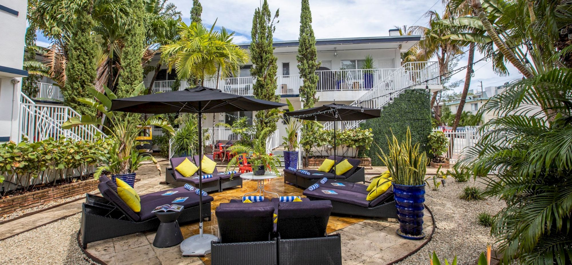 A stylish outdoor lounge area with black wicker seating, yellow cushions, shaded by umbrellas, surrounded by tropical plants and a modern white building.