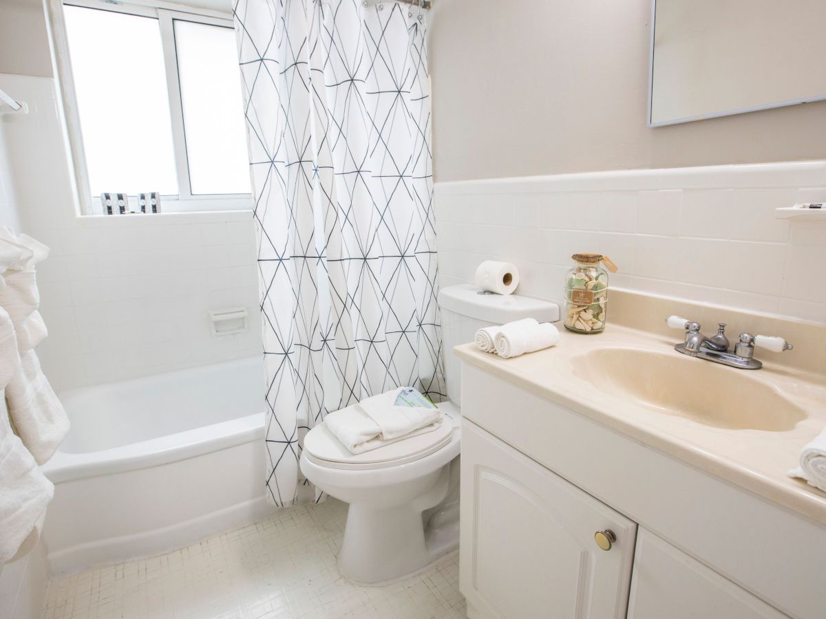 A clean bathroom with a bathtub, shower curtain, toilet, sink, and mirror. Towels and toiletries are neatly arranged on the counter.