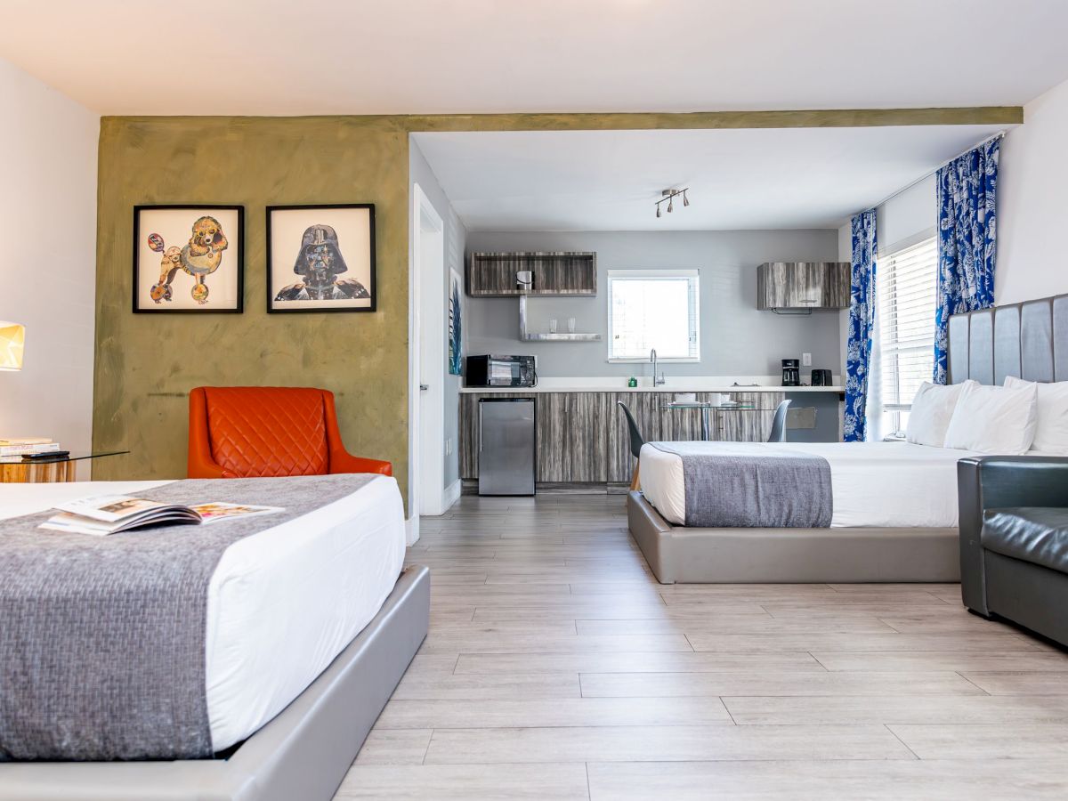 The image shows a modern hotel room with two beds, a kitchenette, couch, and wall art, featuring a contemporary design and cozy atmosphere.