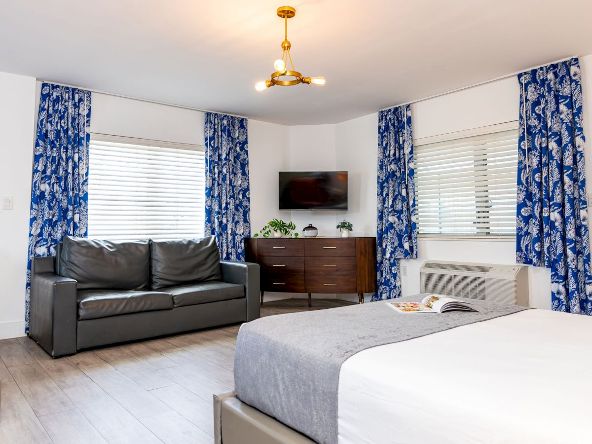 A modern hotel room features a bed, sofa, TV, dresser, and blue curtains. It has a cozy and minimalistic design.