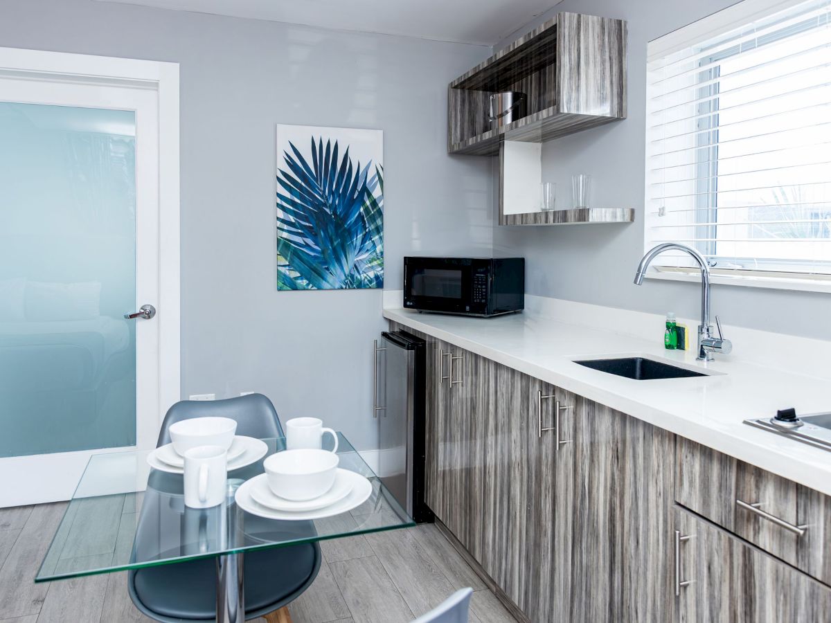 A modern kitchen with wood cabinets, a glass table set with dishes, a microwave, and a leafy wall art, under natural light.