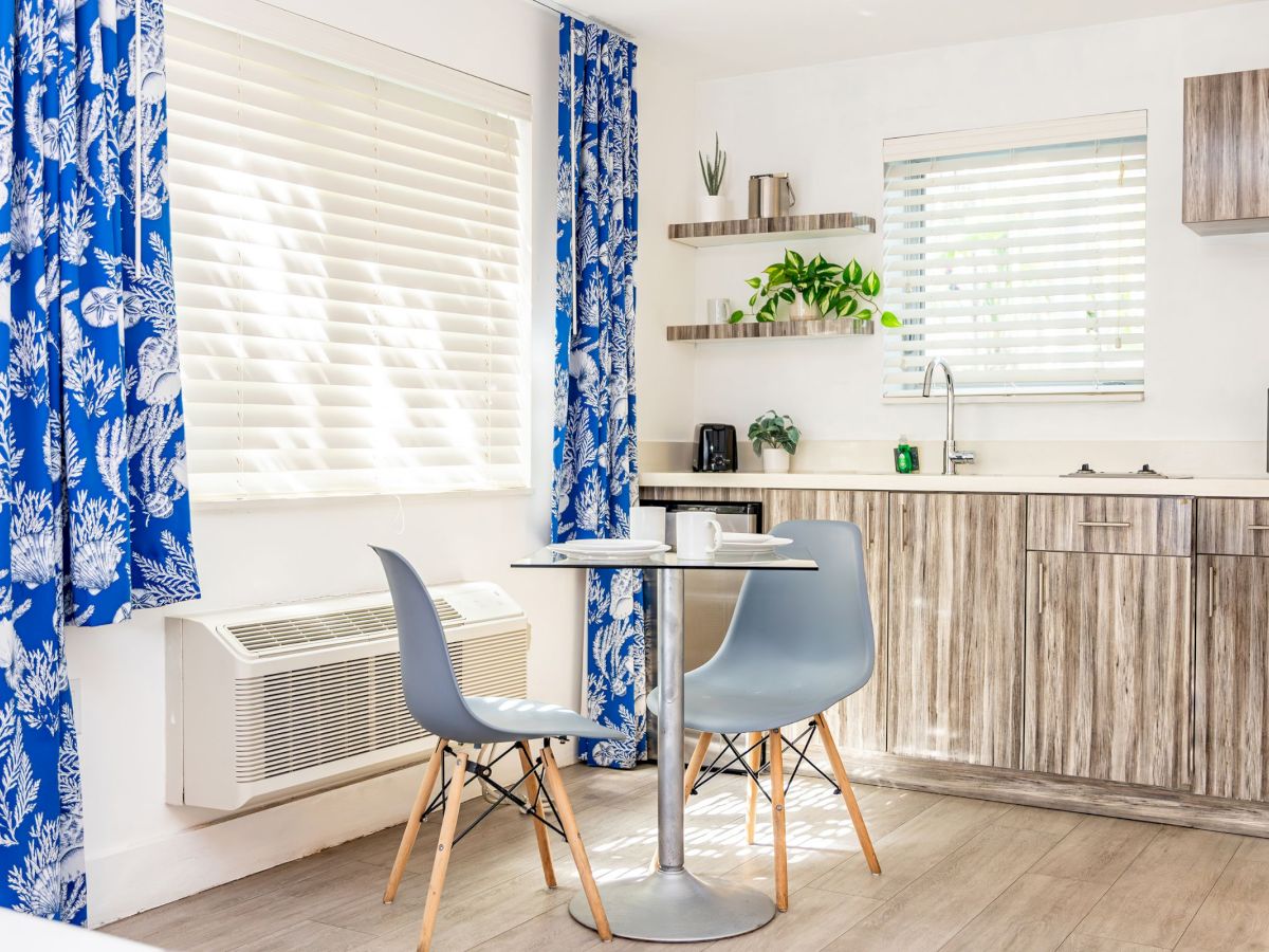 A modern kitchen with blue curtains, a small table with two chairs, wooden cabinets, and greenery, creating a bright and inviting space.
