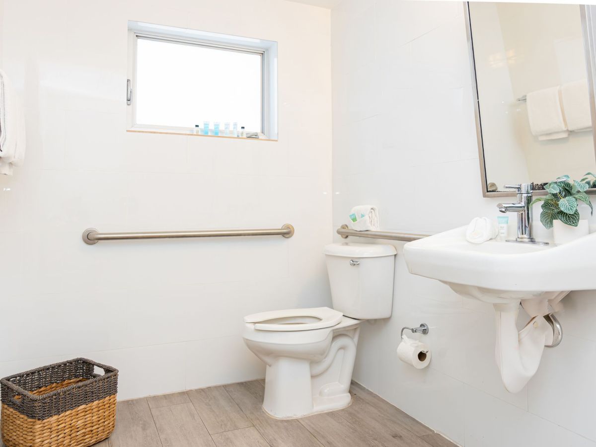 The image shows a clean bathroom with a toilet, sink, mirror, window, and towel basket, featuring a grab bar on the wall.