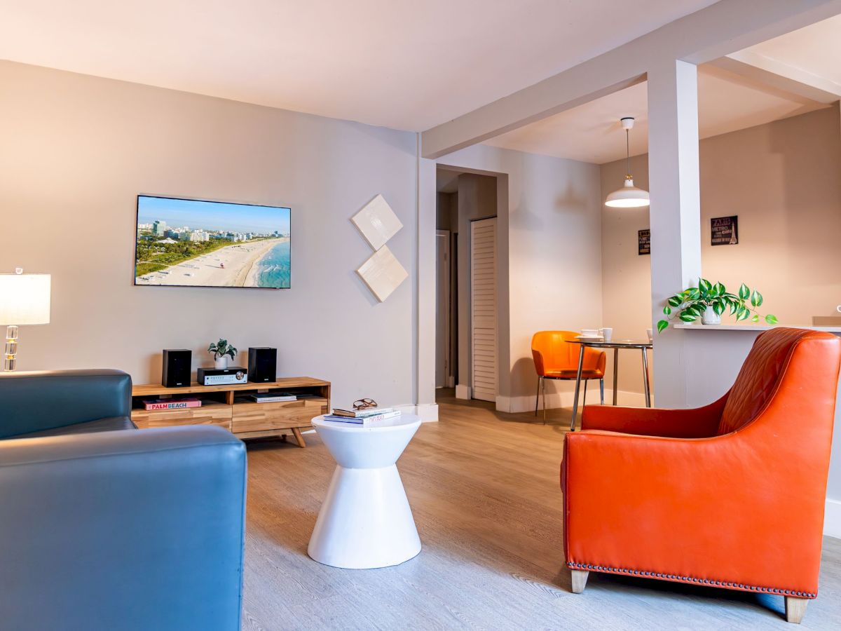 A modern living room with a blue couch, orange chair, TV, and wall art, next to a dining area with a table and chairs, in soft lighting.