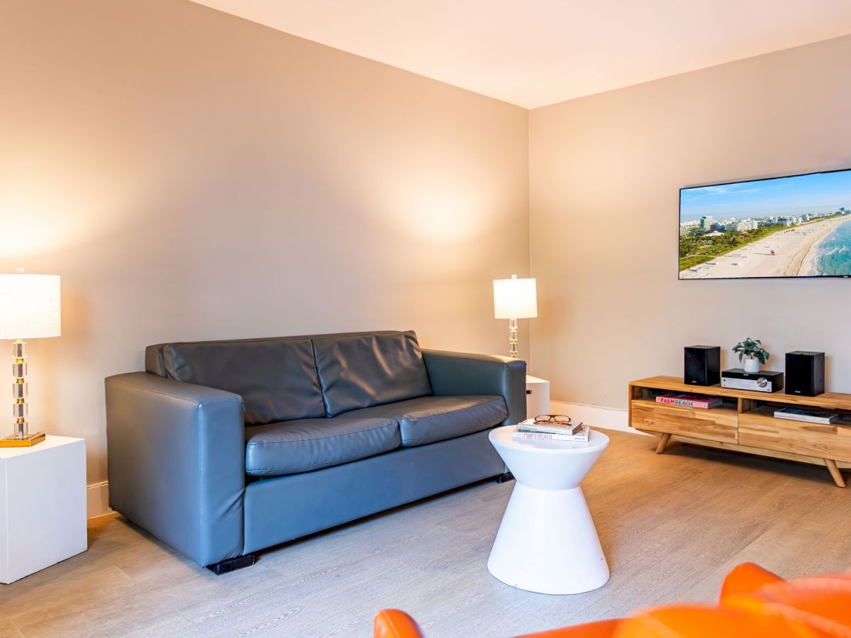 A modern living room featuring a gray sofa, white side tables, two lamps, a wall-mounted TV, and a wooden media console.
