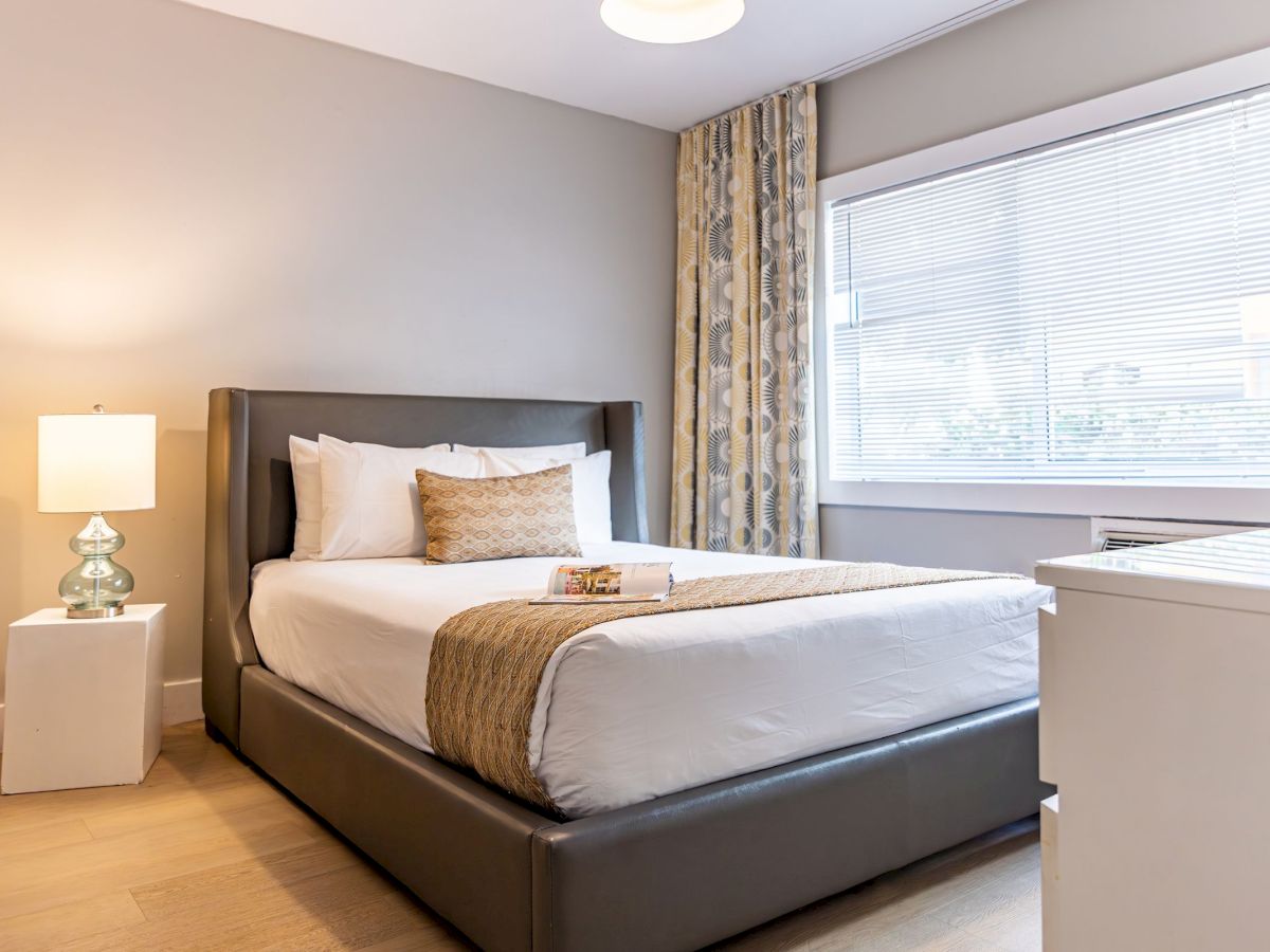 The image shows a tidy bedroom with a neatly made bed, pillows, a lamp on a nightstand, and a large window with curtains.