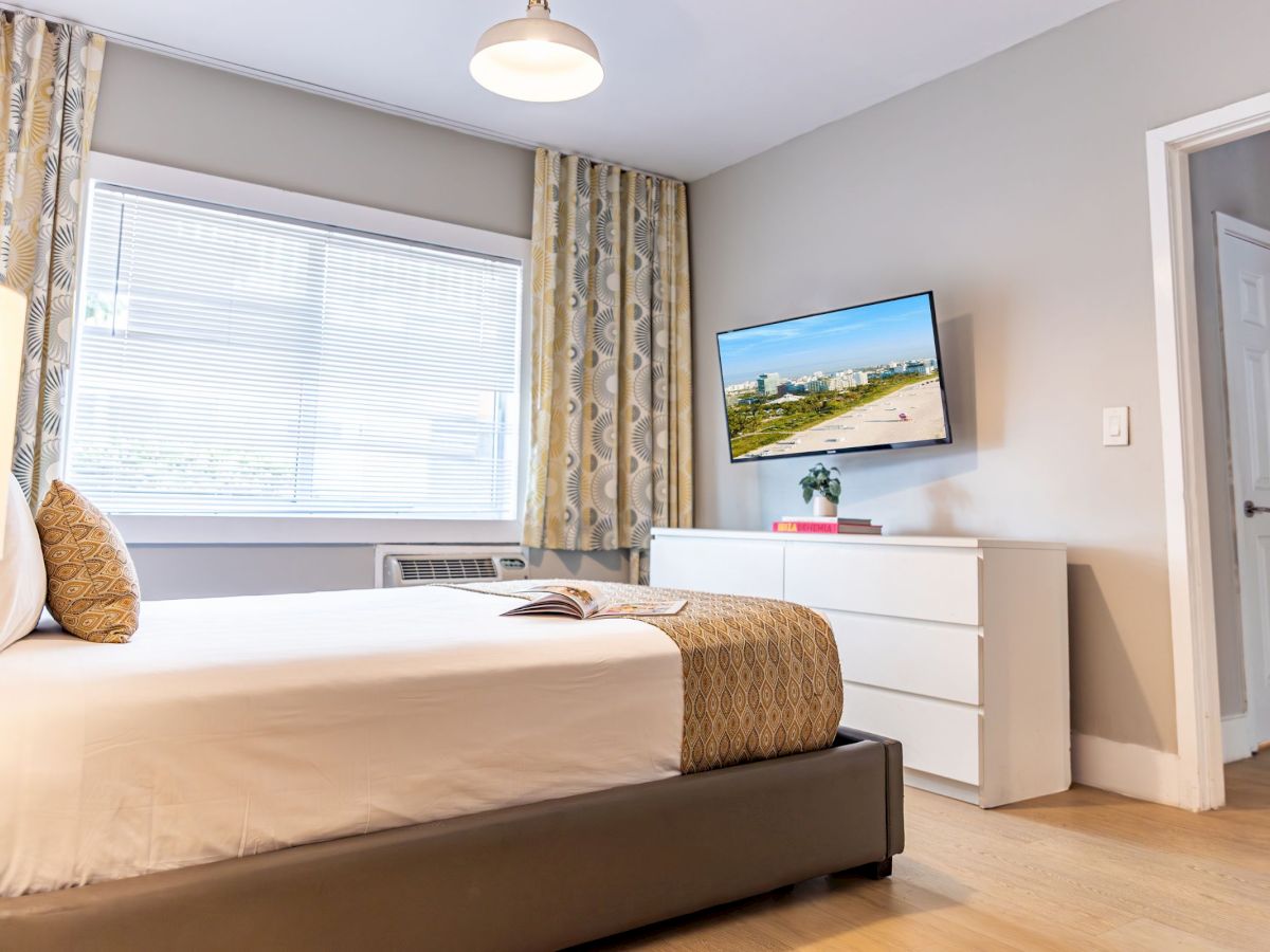 A modern bedroom with a bed, dresser, wall-mounted TV, and window with curtains, all in neutral tones and simple decor.