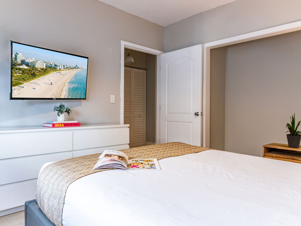 A tidy bedroom with a TV, dresser, plant, and open magazine on a neatly made bed, featuring a modern and minimalistic design.