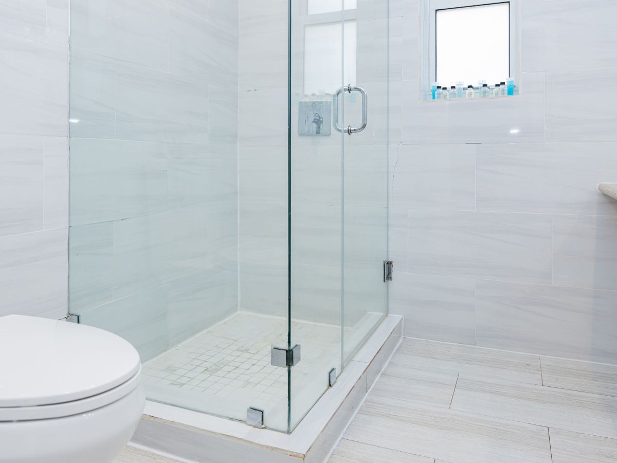 The image shows a modern bathroom with a glass shower, white tiles, and a toilet, creating a clean and minimalist aesthetic.