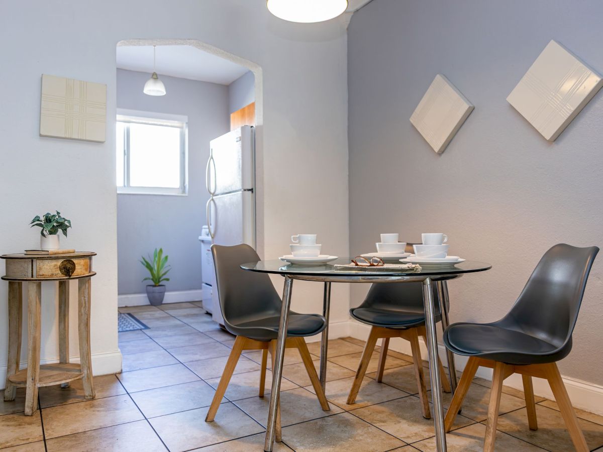 A modern dining area with a round table, four chairs, and tableware, next to a small kitchen, framed by minimalistic wall decor.