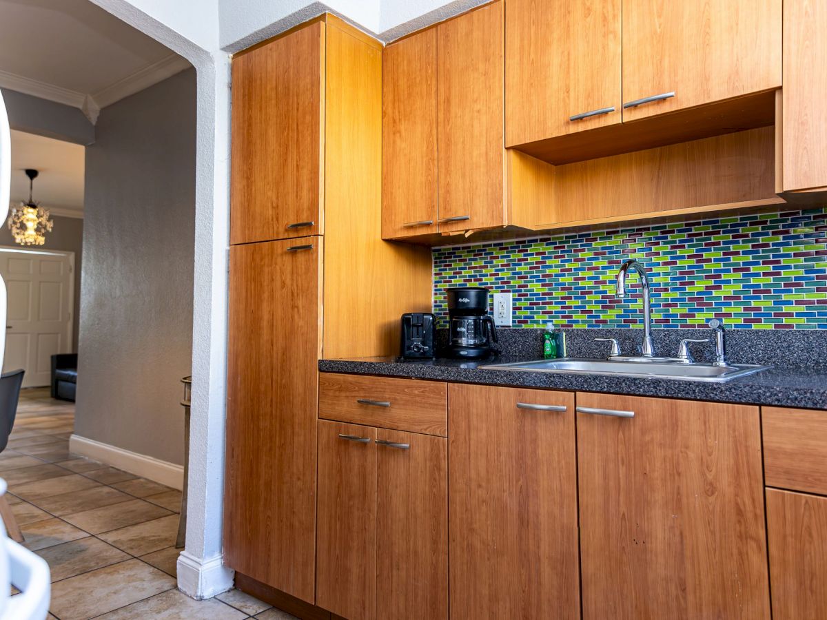 The image shows a kitchen with wooden cabinets, a colorful tiled backsplash, and a coffee maker on the countertop near a sink.