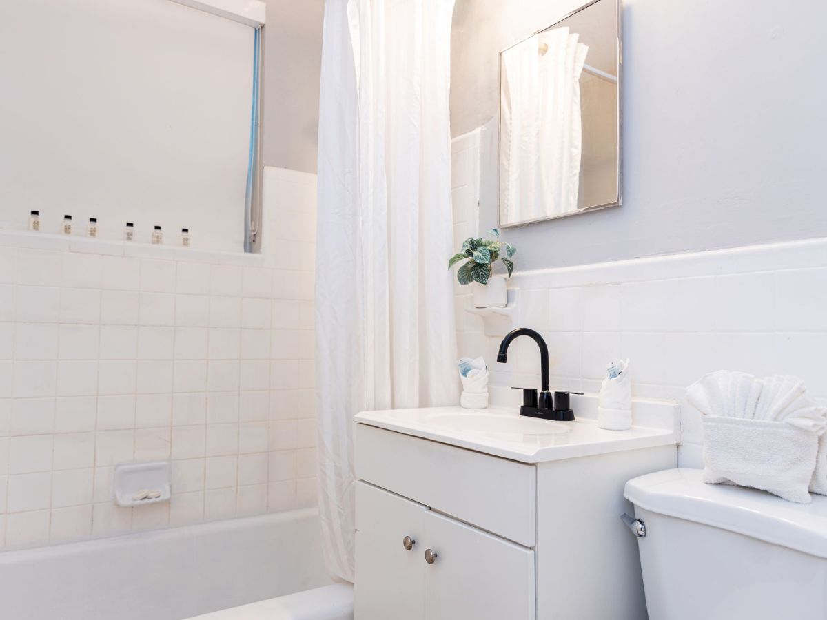 The image shows a minimalist bathroom with a bathtub, a white vanity with a black faucet, a mirror, and a toilet with paper holder art.