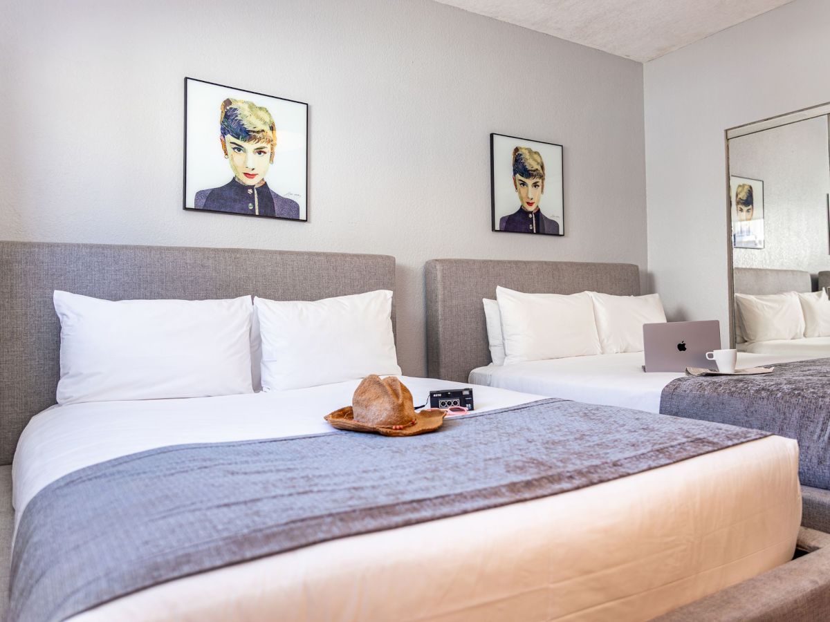 The image shows a modern hotel room with two beds, gray accents, wall art, and a laptop on the bed.