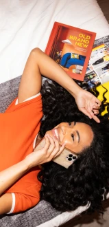 A person in an orange outfit lies on a bed, talking on the phone beside colorful books.