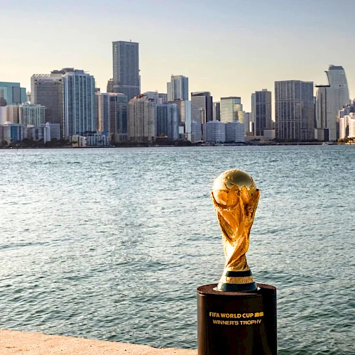 World&rsquo;s largest trophy on a pier by a sparkling harbor with a city skyline in the background, shimmering water, and a clear sky.