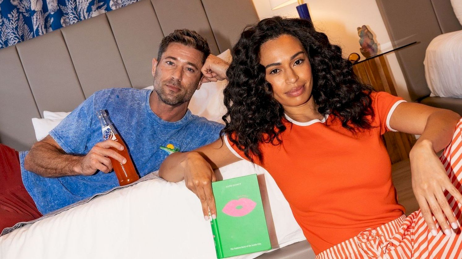 Two people lounge on a bed in a bright hotel room, one in a blue shirt and the other in orange striped pajamas, with sneakers on the floor and a green box between them.