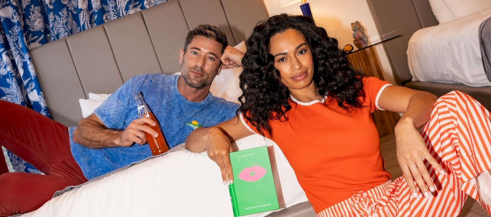 Two people lounge on a bed in a bright hotel room, one in a blue shirt and the other in orange striped pajamas, with sneakers on the floor and a green box between them.