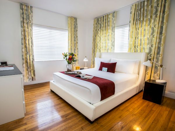 A bright hotel bedroom with a white upholstered bed, red accents, yellow patterned curtains, wooden floors, two bedside tables, and a vase of flowers on the dresser, cozy and modern.