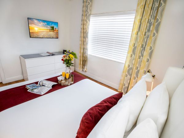 A bright hotel room with a neatly made bed, white linens and burgundy accents, a dresser with a vase of flowers, a wall-mounted TV, and a window with patterned curtains.