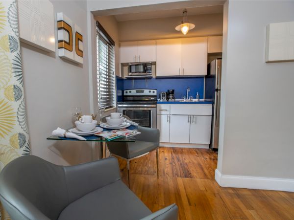 A compact kitchen dining area with a small round table set for two, a gray chair, light wood floors, white cabinets, blue backsplash, and a doorway to a bright kitchen.
