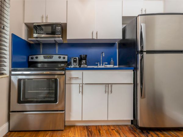 A compact kitchen with stainless appliances: a fridge, stove/oven, microwave, small sink, white cabinets, blue backsplash, and hardwood floors.