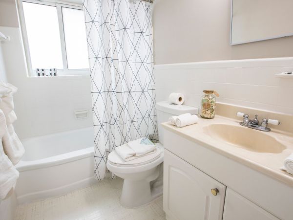 A small bathroom with a bathtub/shower, toilet, and sink. White cabinets, beige countertop, light tiles, and a geometric shower curtain. A few towels and toiletries are visible.