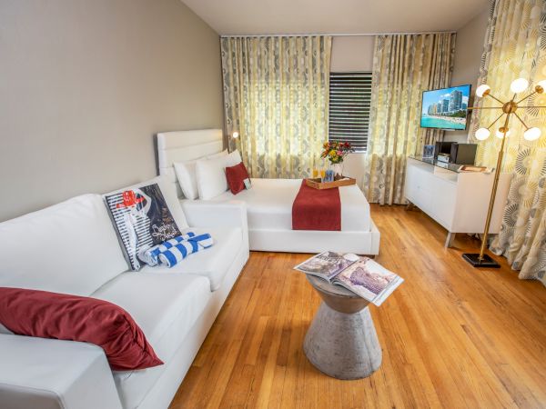 A bright, modern living room with white sofas, red accents, a small round coffee table, and a TV console under patterned curtains.