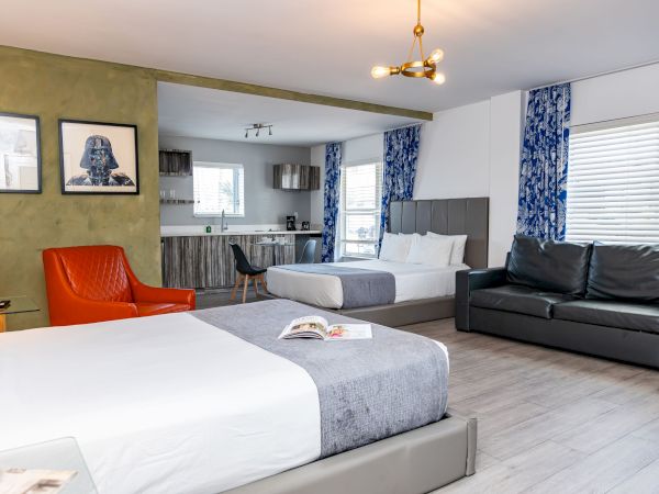 A modern hotel suite with two beds, a red accent chair, a black sofa, a small dining area, and a bright kitchenette in the background, all in a airy, minimalist style.