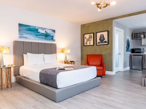 A bright hotel room with a king bed, gray upholstered headboard, two nightstands, a red armchair, artwork, and a small sitting/kitchen area in the background.