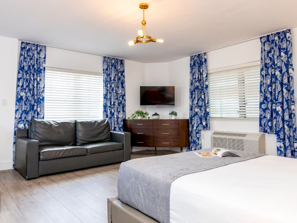 A modern hotel-style suite with a double bed, a dark sofa, a dresser with a TV, blue patterned curtains, and large windows.