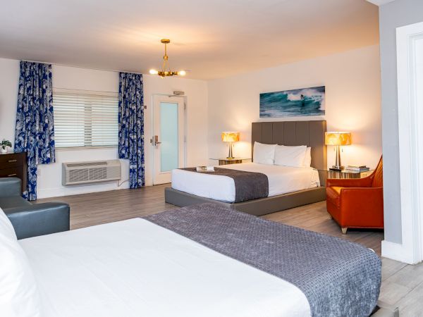 A bright hotel room with two beds, blue curtains, wall art, nightstands, a sofa, and an orange chair, plus a door and window AC unit.