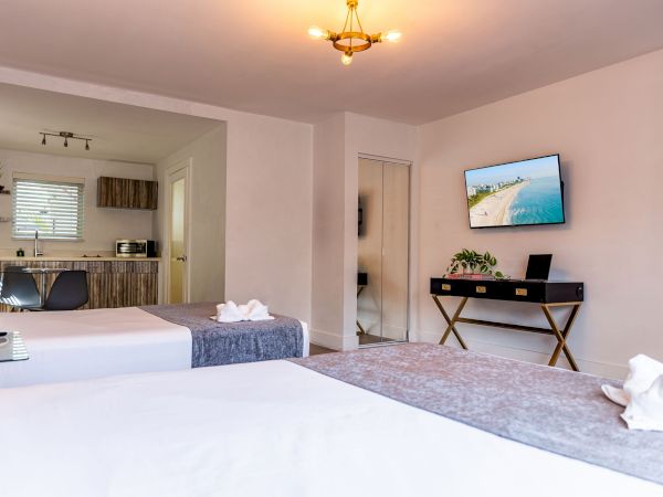 A bright hotel room with two beds, a small desk and TV on the wall, an open doorway to a kitchenette area, and towels arranged on the beds.