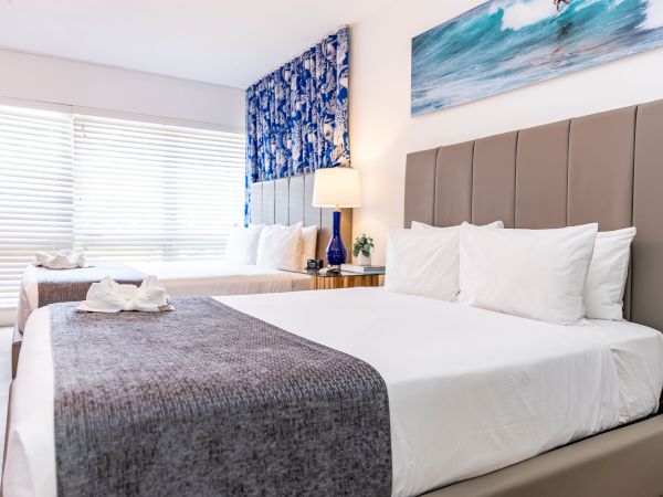 A modern hotel room with two large beds, neutral decor, white linens, a gray bed runner, a nightstand lamp, and ocean art above the headboards.